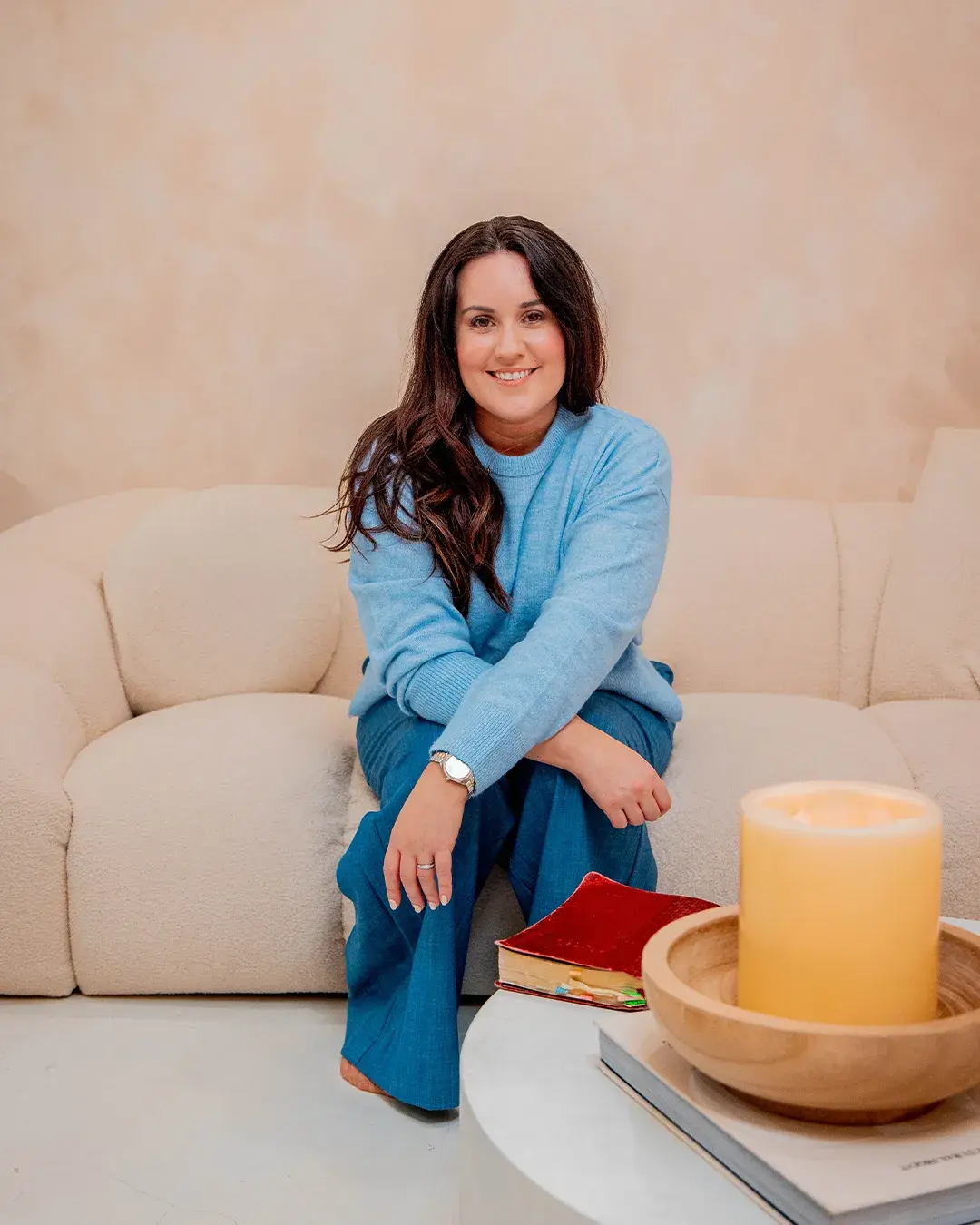 Beth Hobbs sitting on a sofa in a pale blue jumper, smiling, with a candle and a book beside her